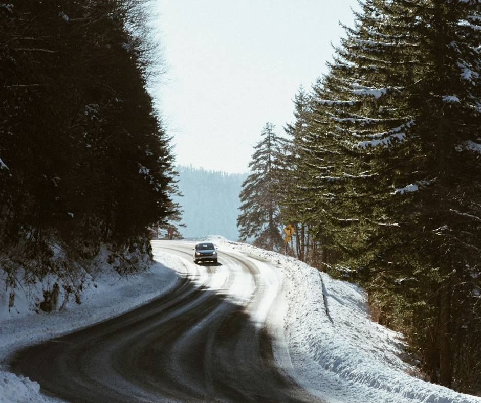 A silver SUV driving along an icy, winding road surrounded by tall trees in winter.