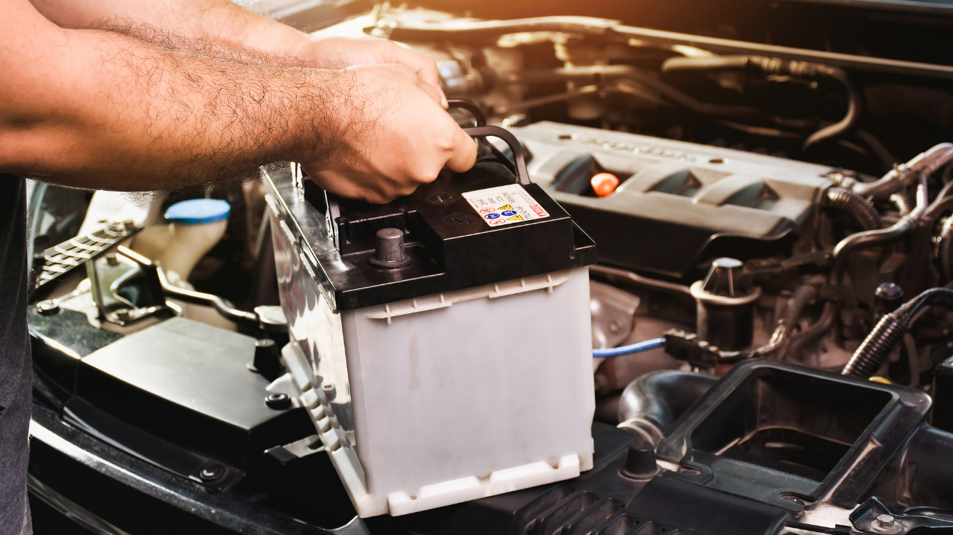 A mechanic lifting a car battery out of an engine compartment during seasonal maintenance.